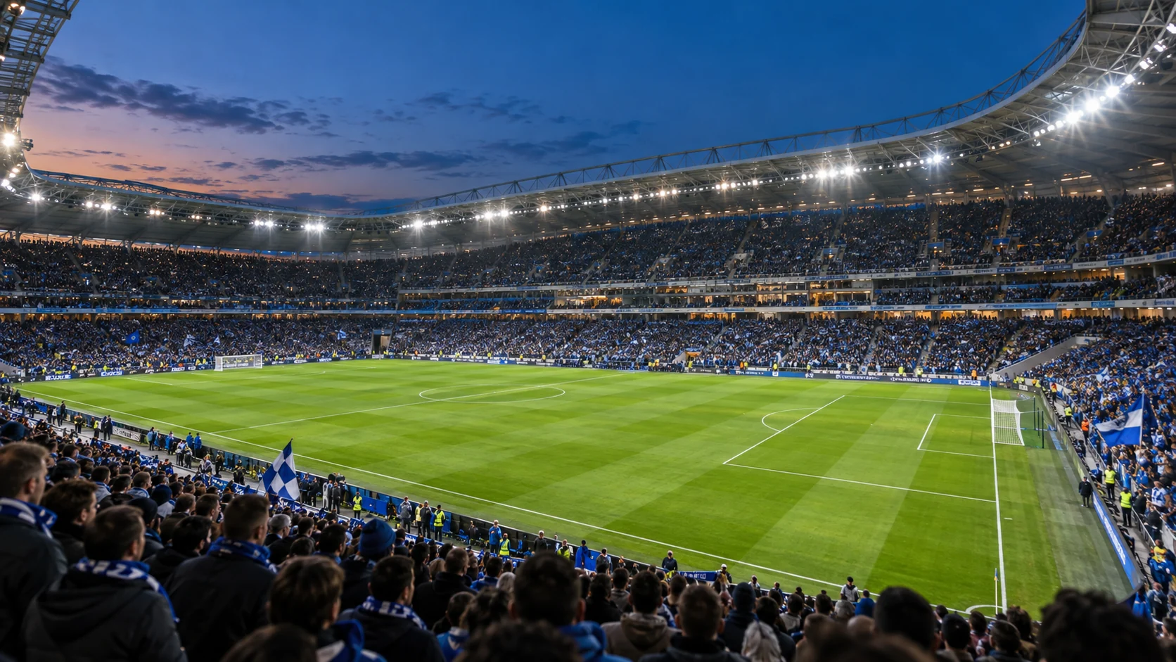 Supporters fill a modern football stadium as floodlights illuminate the pitch at dusk.